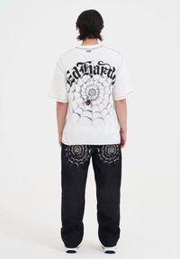Hvid oversized T-shirt med edderkoppedesign og tekst på ryggen, parret med sorte bukser med edderkoppedestillinger.