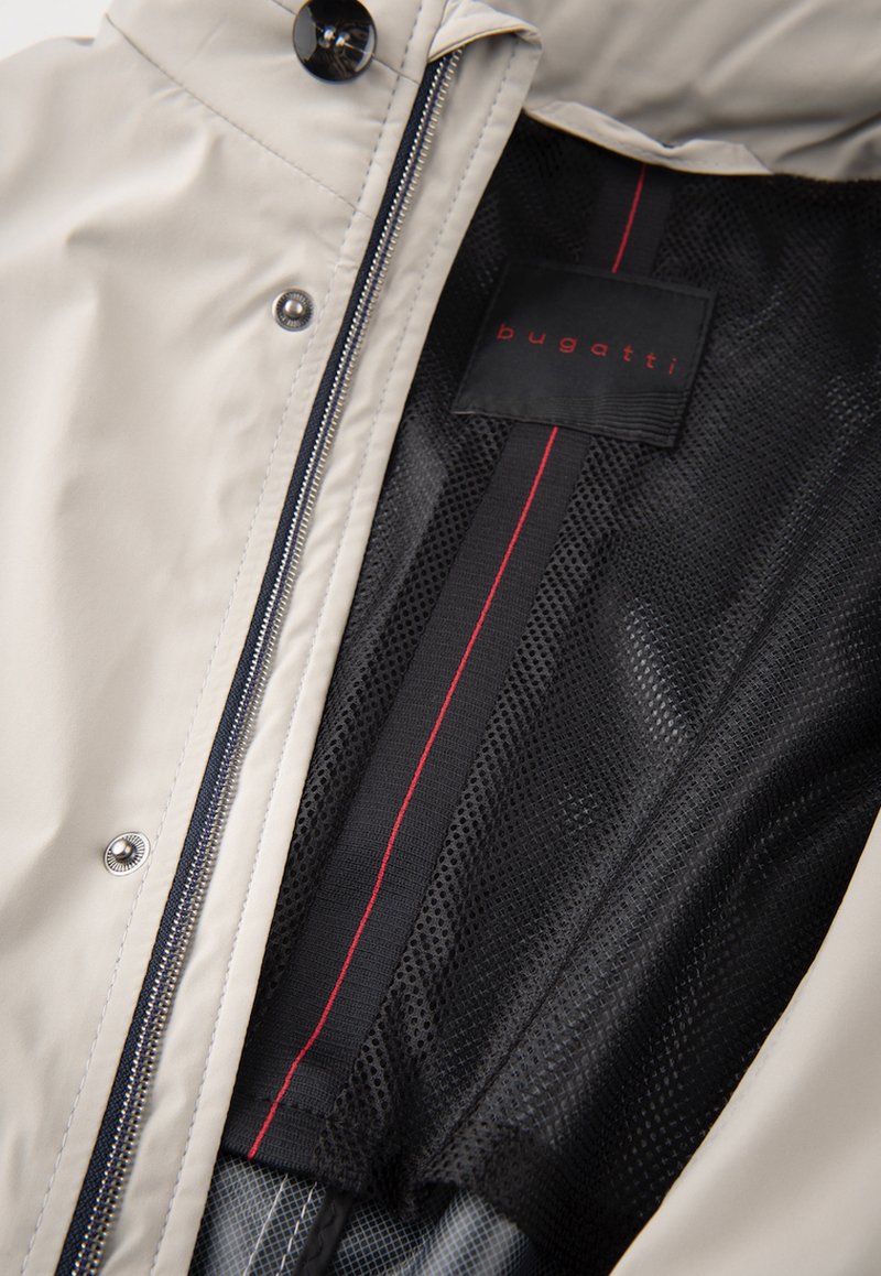 Veste beige clair avec fermeture éclair, dotée d'une doublure en maille noire avec une bande rouge. Étiquette logo affichée en noir à l'intérieur.