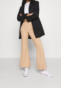 Mujer vestida con pantalones acampanados de talle alto beige, zapatillas altas blancas, camisa blanca, abrigo negro y collar de cadena dorada, de pie con los brazos cruzados.