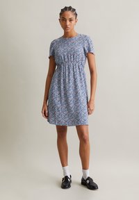 Blumenmusterkleid mit kurzen Ärmeln, runder Halsline und geraffter Taille in Blau, Pink und Weiß. Getragen mit schwarzen Schuhen und weißen Socken.