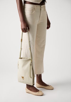 Bolso de cuero beige con cierre de cuerda, acentos en hardware dorado y un acabado texturizado, llevado con zapatos planos beige a juego y pantalones claros.