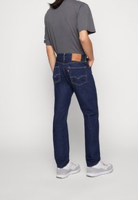 Dunkelblaue Jeans mit geradem Schnitt, ausgestattet mit Gesäßtaschen und einem hellbraunen Lederpatch. Kombiniert mit einem grauen T-Shirt und weißen Sneakers.