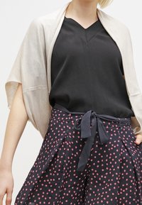 Beżowy sweter z otwartym splotem na czarnej bluzce z dekoltem w serek, w połączeniu z granatowymi szortami w różowe kropki i akcentem związanego pasa.