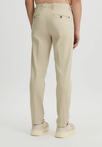 Pantalones beiges ajustados con bolsillos laterales y traseros, con detalles de costura sutiles y una textura suave, combinados con zapatillas de color claro.