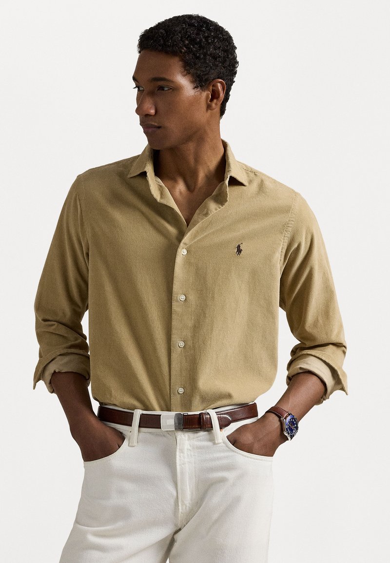 Khaki knappeskjorte med en subtil teksturert mønster, lange ermer og en liten logo, kombinert med hvite bukser og et brunt belte.