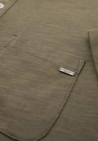 Camicia verde oliva con tessuto testurizzato, tasca frontale e un'etichetta metallica con la scritta "OMBRE DEPT" incisa, caratterizzata da cuciture robuste.