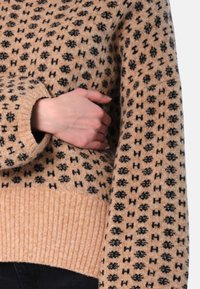 Stickad tröja i beige med svart geometriskt mönster. Har ribbad fåll och muddar, med en avslappnad passform och strukturerad yta.