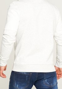 Hombre vestido con un sudadera gris claro y unos jeans oscurecidos de color azul, mostrado desde la espalda con los brazos ligeramente doblados.