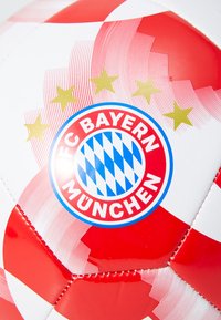 Futbolo kamuolys su blizgiu baltu ir raudonu dizainu, puošiamas FC Bayern München logotipu ir mėlynu languotu raštu su keturiomis auksinėmis žvaigždėmis viršuje.