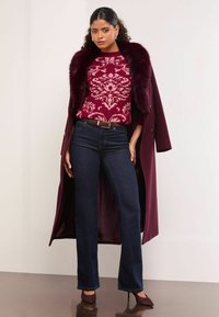 Un palton burgund cu guler din blană sintetică, un pulover cu model floral, blugi cu evazare din denim închis și pantofi cu vârf ascuțit formează o ținută în straturi.