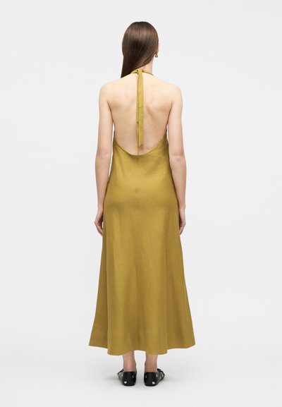 Γυναίκα με μακριά καστανά μαλλιά που φοράει μίντι φόρεμα μουσταρδί με λαιμόκοψη halter στο πίσω μέρος και μαύρα επίπεδα παπούτσια, στέκεται μπροστά σε λευκό φόντο.