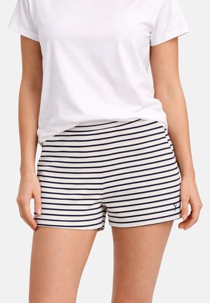 Nainen, joka pukeutuu valkoiseen pyöreäkaulaiseen t-paitaan ja kermaan värillisiin shortseihin, joissa on vaakasuorat laivastonsiniset raidat, seisoo kädet rentoina sivuilla.