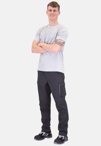 Graues T-Shirt, schwarze Cargo-Hosen mit Seitentaschen und schwarze Sneakers mit grauen und orangefarbenen Akzenten. Das Modell steht mit verschränkten Armen.