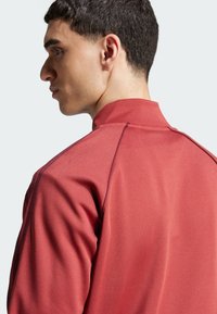 Rote Sportjacke mit hohem Kragen, glatter Oberfläche und flachen Nähten. Sichtbare Schulternahtdetails verleihen dem Design Dimension.