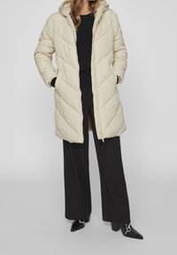 Woman wearing a beige quilted long puffer jacket, black sweater, wide-leg black pants, and pointed black boots, standing with hands in pockets.