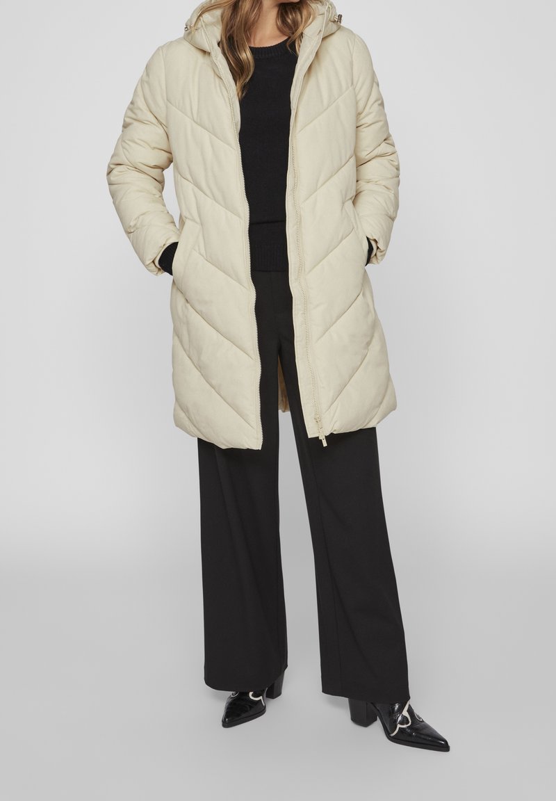 Woman wearing a beige quilted long puffer jacket, black sweater, wide-leg black pants, and pointed black boots, standing with hands in pockets.