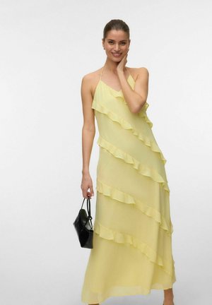 Femme en robe maxi jaune sans manches à volants, tenant un petit sac à main noir, souriante, les cheveux tirés en arrière sur un fond uni.