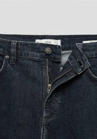 Jean en denim foncé avec une fermeture éclair, un bouton métallique et des coutures apparentes. Le tissu a une texture lisse avec une légère décoloration.