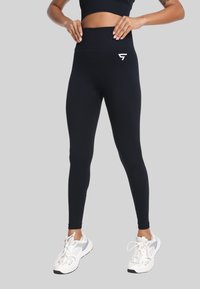 Kobieta w czarnych legginsach z wysokim stanem i białych butach sportowych, stojąca z dłońmi opartymi na biodrach na jednolitym szarym tle.