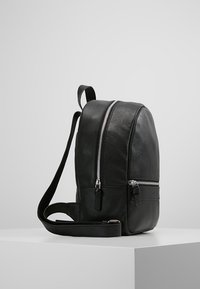 Svart läderryggsäck med två silverfärgade dragkedjor och justerbara remmar, placerad på en vit pedestals mot en enkel grå bakgrund.