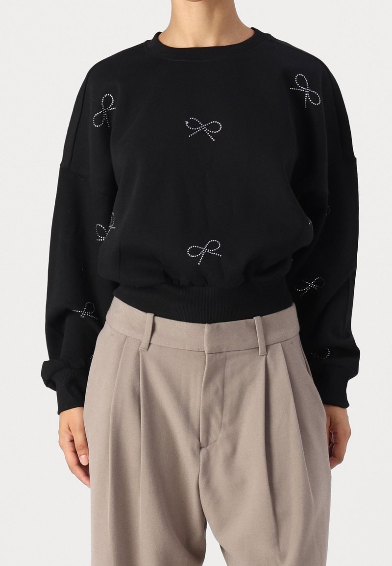Personne portant un sweat-shirt noir à manches longues avec de petits motifs de nœuds blancs à points, associé à un pantalon beige plissé taille haute.