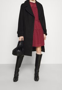 Schwarzer Woll-Trenchcoat mit großen Revers, über einem strukturierten burgunderfarbenen Kleid getragen. Kombiniert mit knielangen schwarzen Lederstiefeln und einer kleinen schwarzen Handtasche.