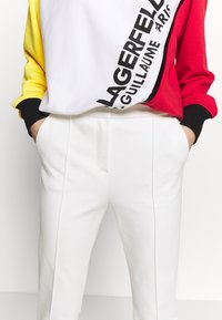 Farbblockiertes Sweatshirt in Weiß, Gelb und Rot mit schwarzen Akzenten und Markenlogo; kombiniert mit enger geschnittener weißer Hose mit Seitennähten.