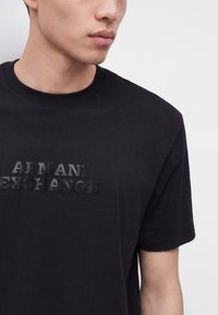 Jeune personne portant un T-shirt noir à manches courtes avec du texte "Armani Exchange" texturé sur le devant, légèrement tournée vers la gauche.