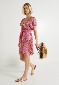 Vestido colorido y estampado con mangas cortas y escote en V, ceñido a la cintura con un cinturón. Combinado con sandalias planas metálicas y un bolso de mimbre.