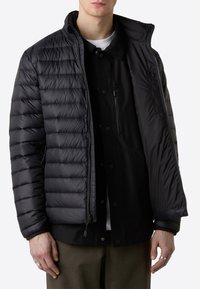 Giacca piumino nera con texture trapuntata, zipper completo e colletto alto. Presenta una tasca laterale e un design a strati su una camicia.
