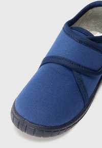 Tumši zila čība, kas izgatavota no teksturēta auduma ar Velcro siksnu. Iekšpusē ir mīksts polsterējums, bet zole ir melna gumija ar rakstainu protektoru.