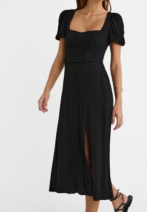 Zwarte midi-jurk met een gestructureerde stof. Heeft pofmouwen en een vierkante halslijn. Voorzien van een zijsplit voor extra detail.