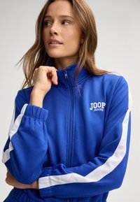 Giacca blu con zip realizzata in tessuto liscio, caratterizzata da strisce laterali bianche e un logo sul petto. Include polsini elasticizzati e colletto alto.