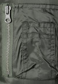Tissu vert avec une fermeture éclair le long du bord gauche, petite poche avec des motifs cousus et une surface texturée. Matériau durable avec une finition mate.