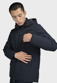 Námořnická voděodolná bunda s kapucí, se zipovými kapsami a hladkým povrchem. Design zahrnuje nastavitelné šňůrky u krku.