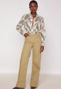 Musterbluse mit langen Ärmeln in hellem Creme mit grünem Blattdesign, kombiniert mit hoch taillierten khakifarbenen weiten Hosen. Mit Knopfverschluss.