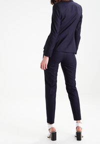 Costume bleu marine sur mesure avec une veste ajustée et un pantalon fuselé, fabriqué en tissu lisse. Présente un design épuré et des accents matériels minimaux.