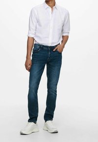 Jeans azul oscuro de corte ajustado, combinados con una camisa blanca de botonadura. Zapatillas blancas completan el conjunto, con un acabado texturizado.