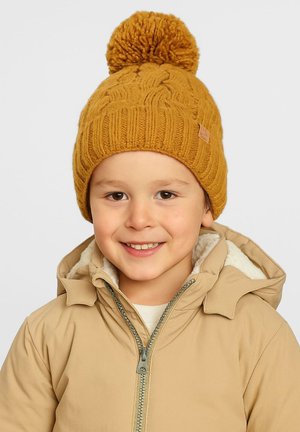 Gorro de lana amarillo con un gran pompón en la parte superior, con un patrón de canalé texturizado. Llevado por un niño con una chaqueta beige.
