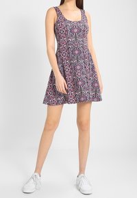 Ärmelloses gemustertes Kleid mit einem pink- und schwarzfarbenen geometrischen Design, tailliertem Oberteil und weit schwingendem Rock, kombiniert mit weißen Sneakers.