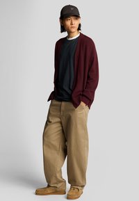 Cardigan bordeaux sopra una maglietta nera, pantaloni beige a gamba larga e scarpe in stile mocassino color cammello, abbinati a un berretto nero e una collana d'oro.