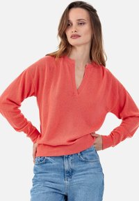 Pull-over corail avec col en V, manches longues et coupe décontractée. Confectionné dans un tissu doux à la texture lisse, associé à un jean bleu clair.