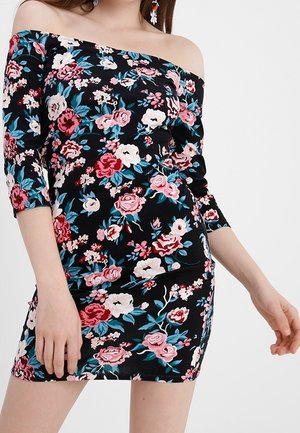 Femme portant une robe noire épaules dénudées avec des motifs floraux roses, rouges, blancs et bleus et des manches trois-quarts.