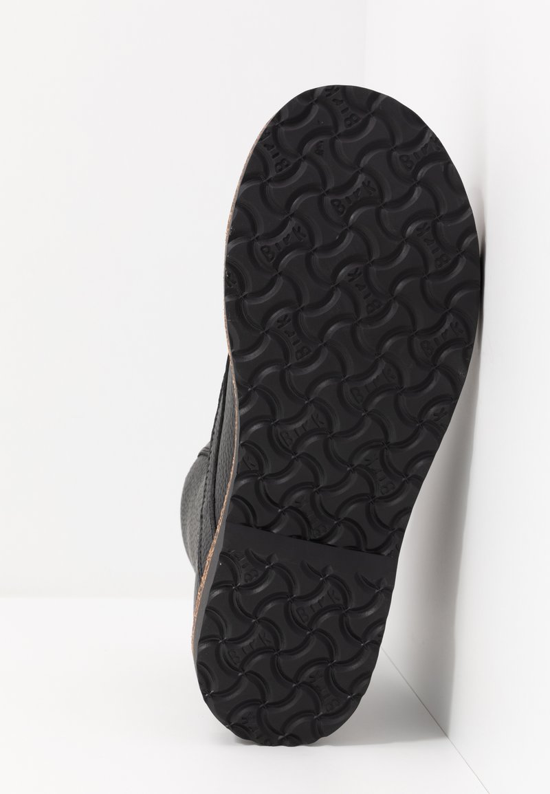 Suola nera di stivale con motivo di battistrada testurizzato e branding "Birk", mostrata su uno sfondo bianco e all'angolo di un muro.