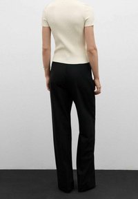 Žena stojící zády k divákovi, oblečená v krémovém krátkým rukávu a volných černých kalhotách na jednobarevném pozadí.