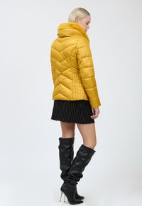 Gul pufferjakke med chevronmønster, der har en krave af falsk pels. Bæres med en sort stribet miniskirt og sort knælang støvler.