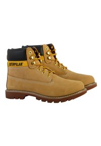 Bottines de randonnée en cuir tan avec des accents noirs, des lacets jaunes et des œillets en métal. Elles possèdent une semelle en caoutchouc robuste et des détails de couture sur les côtés.