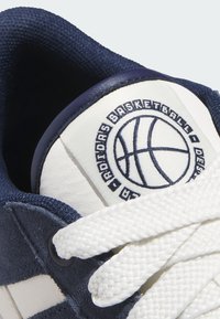 Zbliżenie na język granatowo-białego buta sportowego z logo koszykówki i napisem "Adidas Basketball", z białymi plecionymi sznurówkami.