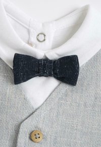 Gilet texturé gris avec un col blanc, featuring une cravate papillon bleu foncé et un détail de bouton en bois. Le design est simple et superposé.