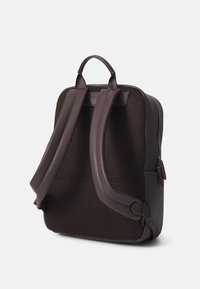 Sac à dos en maille marron avec une surface texturée, des bretelles rembourrées doubles, une poignée sur le dessus et une poche zippée sur le côté pour un rangement supplémentaire.